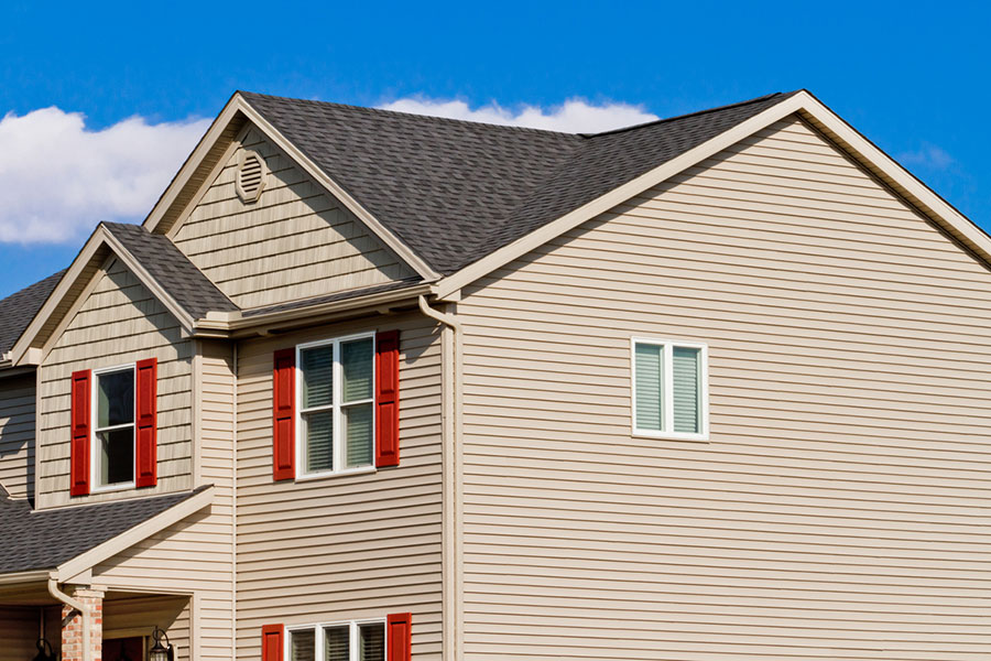 A beige house with gray shingles, red window shutters, and white trim sits under a blue sky with a few clouds, showcasing its fresh look after a recent siding replacement. Perfect for those seeking Michigan home siding inspiration.
