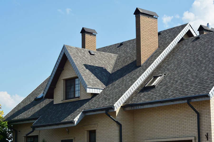 A brick house with a dark gray shingle roof, two chimneys, dormer windows, and white trim is shown under a blue sky with some clouds. Designed to withstand summer heat and help prevent roof damage, the durable materials contribute to an impressive roof lifespan.